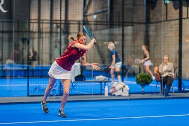 Riga, Letonya - 5 Nisan 2025 - Padel turnuvasında arka planda seyirciler ve diğer oyuncuların yer aldığı kadın padel oyuncusu salıncakta sallanıyor.