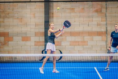 Riga, Letonya - 5 Nisan 2025 - Padel turnuvasında iki kadın yarışıyor, bir oyuncu kalede sallanırken diğeri geride, bir sonraki hamle için hazır..