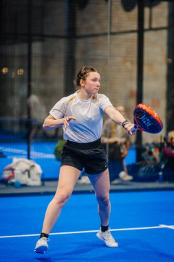Riga, Letonya - 5 Nisan 2025 - Odaklanmış bayan Padel oyuncusu, kapalı alanda oynanan bir maçta ön atış hazırlıyor..