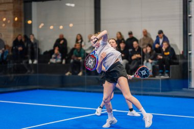 Riga, Letonya - 5 Nisan 2025 - Bir turnuva maçında eşzamanlı çekim yapan iki bayan Padel oyuncusu, arka planda izleyen bir kalabalıkla.