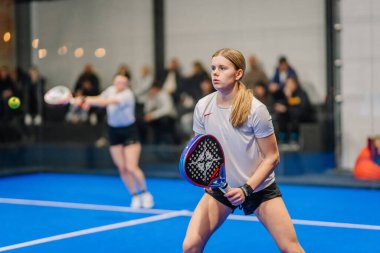 Riga, Letonya - 5 Nisan 2025 - İki bayan oyuncu yoğun bir Padel turnuvasında yarışıyor, biri kalede hazır, diğeri şut atıyor. Seyirciler tribünden izliyor.