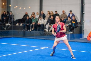 Riga, Letonya - 5 Nisan 2025 - Kırmızı tişörtlü ve beyaz etekli bayan padel oyuncusu, arka planda seyircilerin izlediği bir Padel turnuvası sırasında tekrar atış yapmaya hazırlanıyor..