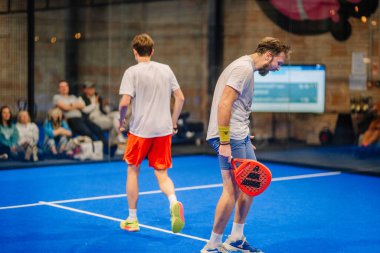 Riga, Letonya - 5 Nisan 2025 - Bir noktadan sonra iki padel oyuncusu, bir puan tablosu ve seyirciler arka planda görünürken bir tanesi hayal kırıklığını ifade ediyor.