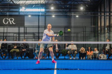 Riga, Letonya - 5 Nisan 2025 - Padel turnuvası sırasında bir erkek Padel oyuncusu topa vurmak için camdan izleyen bir seyirciyle havanın ortasına atlar..