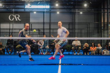 Riga, Letonya - 5 Nisan 2025 - Yoğun bir turnuva maçının ortasında iki erkek padel oyuncusu, seyirci kapalı bir sahanın cam duvarının arkasından yakından izliyor.
