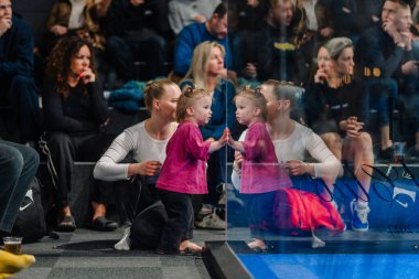 Riga, Letonya - 5 Nisan 2025 - Yürümeye yeni başlayan bir çocuk annesiyle Padel Turnuvası 'nı izlerken cam bir duvara dokunur ve kalabalık bir ev ortamında güzel bir yansıma anı yaratır..