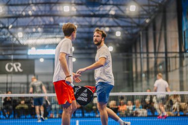 Riga, Letonya - 5 Nisan 2025 - Padel turnuvasında arka planda seyirciler ve diğer oyuncularla oynanan maçta iki erkek Padel oyuncusu sahada etkileşime girer.