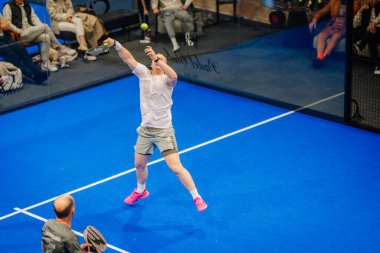 Riga, Letonya - 5 Nisan 2025 - Erkek Padel oyuncusu, kapalı bir padel turnuvasında oynanan yoğun bir maçta güçlü bir atış için atlarken takım arkadaşı arkadan izliyor..