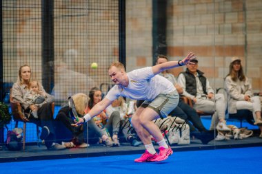 Riga, Letonya - 5 Nisan 2025 - Erkek Padel oyuncusu maç sırasında topa ulaşmak için uzanıyor, odaklanmış seyirciler kapalı bir padel turnuvasında camın arkasından izliyor..