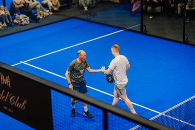 Riga, Letonya - 5 Nisan 2025 - İki erkek Padel oyuncusu, kapalı alanda oynanan bir Padel turnuva sırasında seyircilerin önünde takım çalışmasını kutlayarak sahada beşlik çaktı.