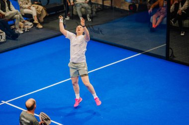 Riga, Letonya - 5 Nisan 2025 - Erkek Padel oyuncusu, kapalı bir padel turnuvasında oynanan yoğun bir maçta güçlü bir atış için atlarken takım arkadaşı arkadan izliyor..