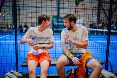 Riga, Letonya - 5 Nisan 2025 - Mola sırasında bir bankta oturan iki erkek padel oyuncusu, ellerinde su şişeleri ve kapalı bir padel turnuvası sırasında sohbet ediyorlar..