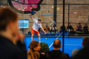Riga, Letonya - 5 Nisan 2025 - Erkek oyuncu bir padel turnuvası maçında servis yapmaya hazırlanırken, seyirciler kapalı sahadaki cam duvarın arkasından izleyecekler..