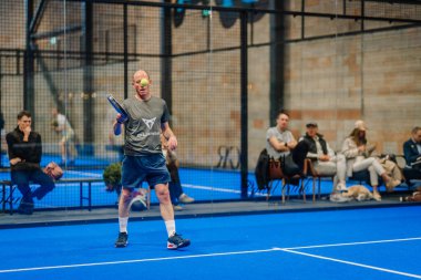 Riga, Letonya - 5 Nisan 2025 - Padel turnuvası maçında top havada ve seyirciler arka planda otururken üst düzey erkek Padel oyuncusu servis yapmaya hazırlanıyor..