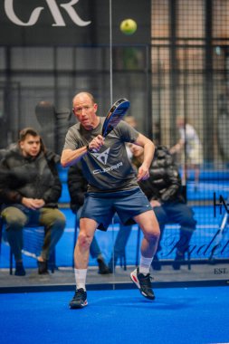 Riga, Letonya - 5 Nisan 2025 - Kapalı alan padel turnuvası sırasında erkek oyuncu, topu geri vermeye odaklandı, seyirciler camın arkasından izliyor..