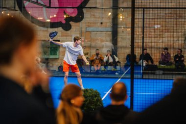 Riga, Letonya - 5 Nisan 2025 - Erkek oyuncu bir padel turnuvası maçında servis yapmaya hazırlanırken, seyirciler kapalı sahadaki cam duvarın arkasından izleyecekler..