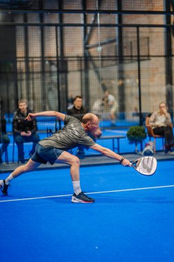 Riga, Letonya - 5 Nisan 2025 - Padel turnuvası maçında arka planda seyircilerin olduğu canlı mavi bir sahada son sınıf erkek Padel oyuncusu topa ulaşmak için geriniyor..