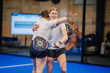 Riga, Letonya - 5 Nisan 2025 - Padel turnuvasını kazandıktan sonra iki genç bayan Padel oyuncusu gülümseyerek ve raketlerini kortta tutarak neşeyle sarılıyorlar..