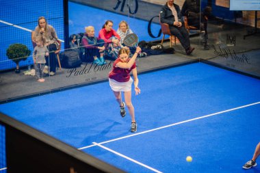 Riga, Letonya - 5 Nisan 2025 - Padel turnuvası sırasında çocuklar ve aileler de dahil olmak üzere seyircilerin saha kenarından izlediği kadın padel oyuncusu topa vuruyor..