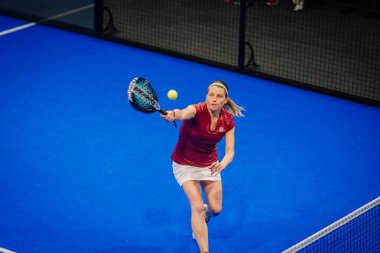 Riga, Letonya - 5 Nisan 2025 - Padel turnuvası sırasında cam duvarların arkasında seyirci varken düşük topa uzanan kadın oyuncu.