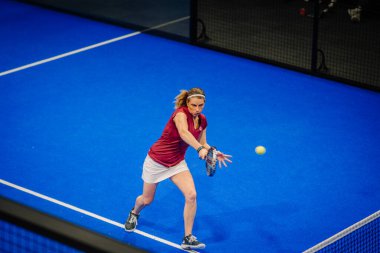 Riga, Letonya - 5 Nisan 2025 - Padel turnuvası sırasında cam duvarların arkasında seyirci varken düşük topa uzanan kadın oyuncu.