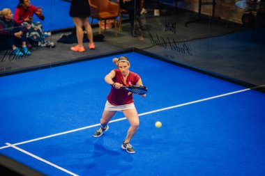 Riga, Letonya - 5 Nisan 2025 - Padel turnuvası sırasında cam duvarların arkasında seyirci varken düşük topa uzanan kadın oyuncu.