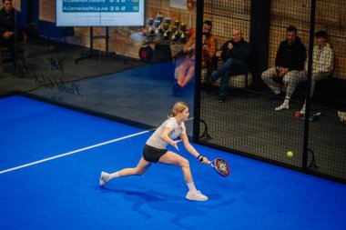 Riga, Letonya - 5 Nisan 2025 - Dişi sporcu, arka planda izleyiciler ve kupalar ile bir padel turnuva maçında düşük bir topu geri vermek için hamle yapar.