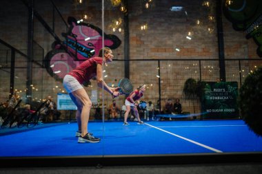 Riga, Letonya - 5 Nisan 2025 - Kırmızı tişörtlü iki bayan oyuncu, canlı bir iç sahada seyircilerle oynanan Padel turnuvası maçında servis almaya hazırlanıyorlar..