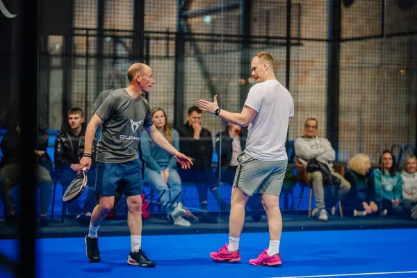 Riga, Letonya - 5 Nisan 2025 - Turnuva maçında iki Padel oyuncusu bir noktayı tartışırken, seyirciler camların arkasından dikkatle izliyor.