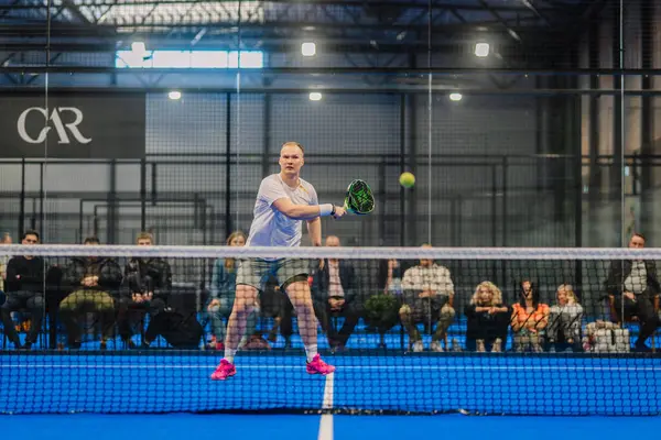 Riga, Letonya - 5 Nisan 2025 - Padel turnuvası sırasında bir erkek Padel oyuncusu topa vurmak için camdan izleyen bir seyirciyle havanın ortasına atlar..