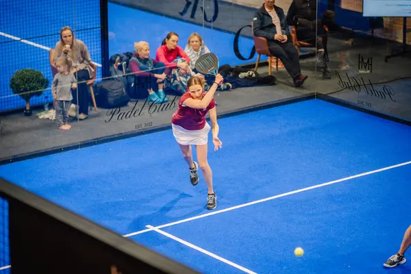 Riga, Letonya - 5 Nisan 2025 - Padel turnuvası sırasında çocuklar ve aileler de dahil olmak üzere seyircilerin saha kenarından izlediği kadın padel oyuncusu topa vuruyor..