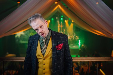 Şık giyinmiş, başı eğik ve komik bir yüz ifadesi olan adam dekoratif perdeleri ve sahne ışıklarıyla zarif bir mekanda poz veriyor..