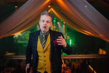 Süslü takım elbiseli bir adam dekoratif perdeleri ve sahne ışıkları olan zarif bir mekanda konuşurken şaşırmış ya da komik bir yüz ifadesi takınıyor..