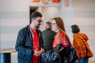 Riga, Letonya - 12 Nisan 2025 - Kırmızı formalı iki florbol taraftarı, turnuva finalleri sırasında, sıradan bir ortamda diğer katılımcılar tarafından çevrelenmiş, kapalı alanlarda konuşuyorlar..