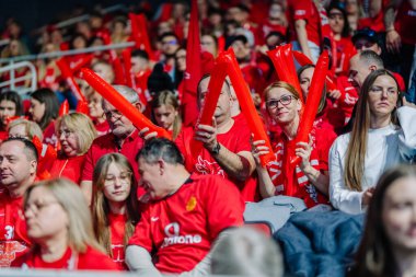 Riga, Letonya - 12 Nisan 2025 - Kırmızı tişörtlü tutkulu fanatikler zemin topu finallerinde şişirilebilir sopalarla tezahürat yapıyorlar, enerjik kalabalık enerjisi, spor etkinliği atmosferi.