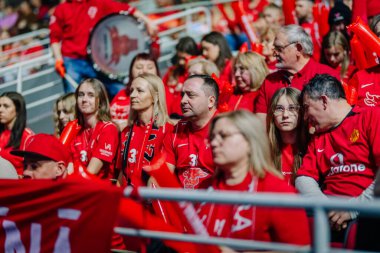 Riga, Letonya - 12 Nisan 2025 - Kırmızı elbiseli fanatikler arenada oturmuş, floorball finalleri boyunca klapa ve eşarp tutuyorlar, takım ruhunu ve desteğini gösteriyorlar..