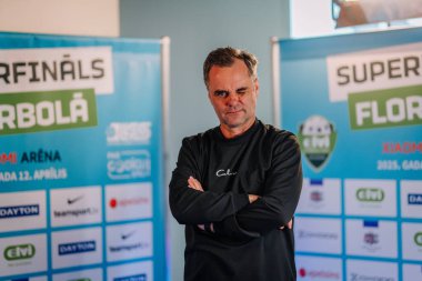 Riga, Letonya - 12 Nisan 2025 - Siyah gömlekli kendinden emin bir adam kolları çapraz pozlar ve sponsor logolarının yer aldığı zemin topu finalleri önünde göz kırpar.