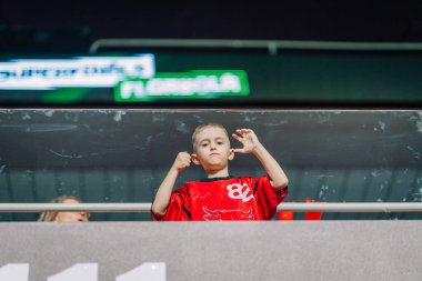 Riga, Letonya - 12 Nisan 2025 - Kırmızı tişörtlü genç bir fanatik salon arenasında, tutku ve takım ruhunu sergileyerek yumruk tokuşturup alkışlıyor.