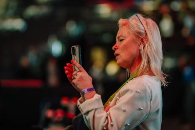 Riga, Letonya - 12 Nisan 2025 - Sarı saçlı bir kadın ev içi bir etkinlikte akıllı telefonuyla fotoğraf veya video çekiyor..