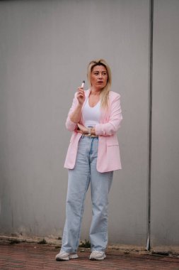Riga, Letonya - 12 Nisan 2025 - Pembe ceketli sarışın kadın, gri bir duvara yaslanırken elinde bir kalem tutuyordu..