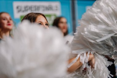 Riga, Letonya - 12 Nisan 2025 - Beyaz ponpon kızlı ponpon kız finallerde sahne alacak, gözleri ve yüzü bulanık arka plan ve diğer sanatçılara odaklan.