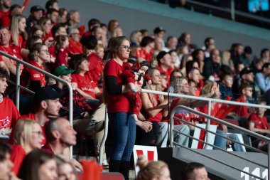 Riga, Letonya - 12 Nisan 2025 - Bir anne, kırmızılı taraftarlarla çevrili bir şekilde florbol finalinde bebeğini kucağına alır ve kalabalıkta aile desteğinin dokunaklı bir anını yakalar..