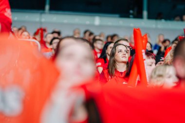 Riga, Letonya - 12 Nisan 2025 - Kırmızı tişörtlü odaklanmış bayan hayran kalabalık bir arenada bayrak ve köpük sopalarla destekçilerle çevrili florbol finallerini izliyor.