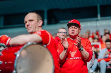 Riga, Letonya - 12 Nisan 2025 - Kırmızı gömlekli ve şapkalı FBK Valmiera taraftarları kalabalık bir arenada tutku ve destek sergileyerek final maçlarında davul çalıyorlar..