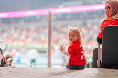 Riga, Letonya - 12 Nisan 2025 - Kırmızı takım tişörtlü küçük bir çocuk yer topu finalleri sırasında arena merdivenlerinde oturuyor, kameraya bakıyor, renkli bulanık arkaplan, spor etkinliği.