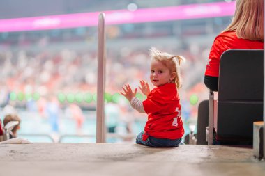 Riga, Letonya - 12 Nisan 2025 - Stadyum merdivenlerinde oturan kırmızı takım tişörtlü küçük çocuk, geriye bakıyor ve gülümsüyor, arka planda bulanık arena ve oyuncular.