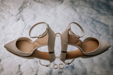 Beyaz gelinlik ayakkabıları ve altın yüzükleriyle zarif bir düğün kompozisyonu yumuşak ışıkla yansıtıcı bir yüzeyde yaratıcı bir şekilde düzenlenmiştir..