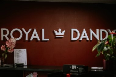 Valmiera, Letonya - 9 Ağustos 2024 - Kraliyet Dandy salonunun resepsiyon alanı kırmızı bir duvarda logo, fiyat listesi, çiçekler ve dekoratif unsurlar ile şık bir iç atmosfer yaratıyor..