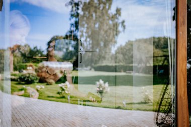 Valmiera, Letonya - 9 Ağustos 2024 - Elinde ayna ve telefon olan bir kadının yansıması, bir düğün sırasında arka planda bahçe manzaralı cam bir pencereden görülüyor..
