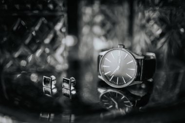 Arka planda bulanık kristal bir cam ile yansıtıcı bir yüzey üzerinde bir kol saati ve kol düğmeleri zarif siyah ve beyaz yakın plan.